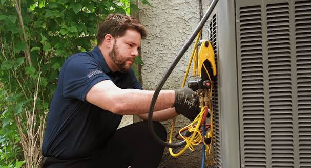 Technician servicing a residential outdoor air conditioner beside garden foliage
